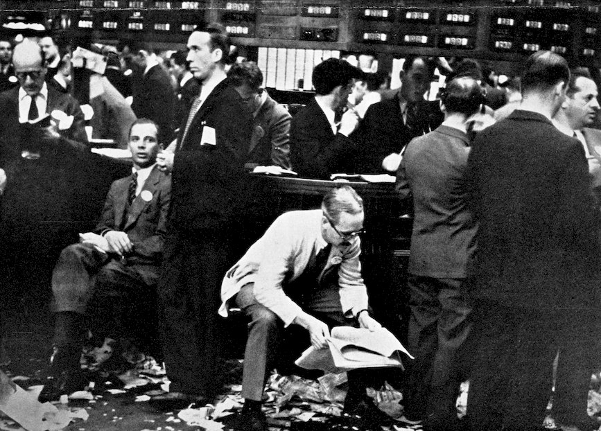 A busy, historical black and white photograph of a stock exchange trading floor, with men in suits reading papers amidst scattered ticker tape on the ground.