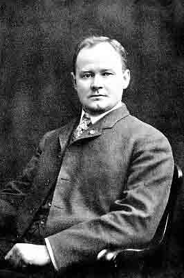 Torso view; four-button suit with vest, windsor tie; seated in wooden chair with scrolled arms.