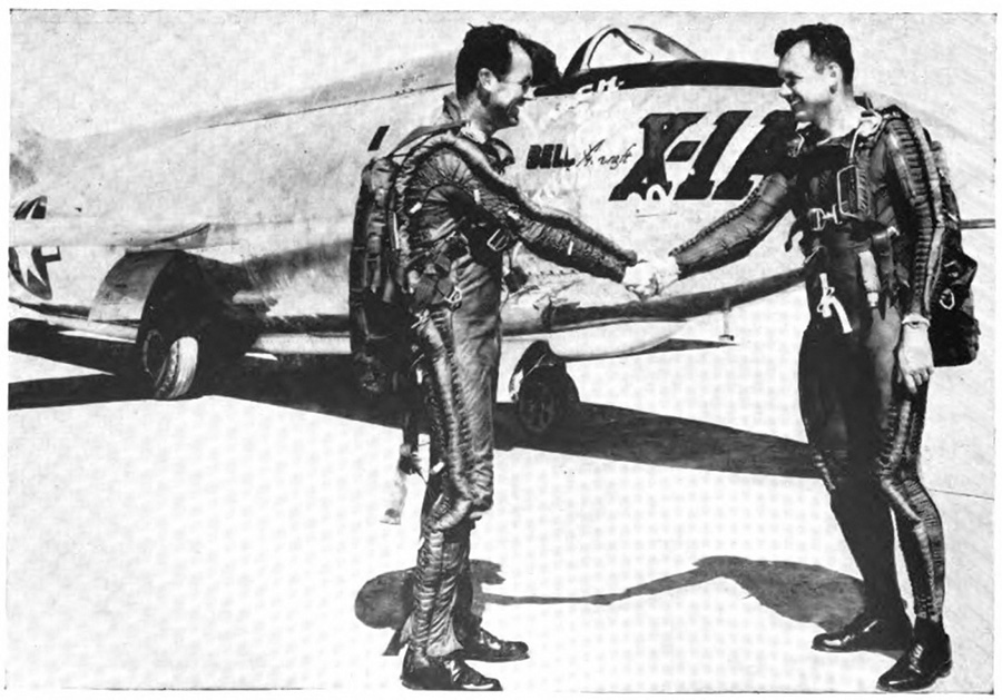 Two men in tightly fitted suits shaking hands in front of an airplane