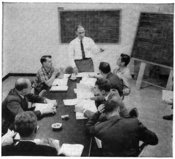 A man standing at the head of a table with people sitting listening to him and papers in front of them 