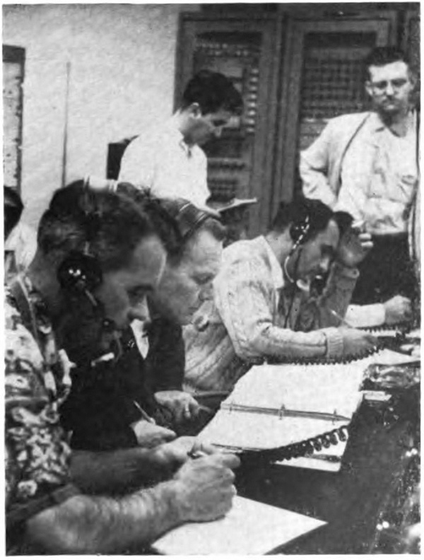 Men sitting at a long table with papers in front of them; a couple of men are standing