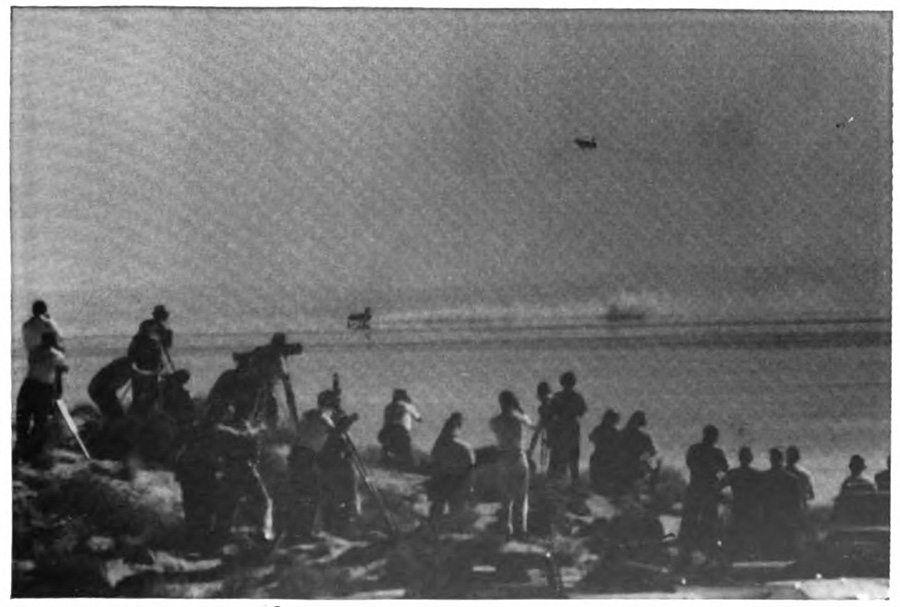 A crowd of people watching a plane in the distance from a lakeshore